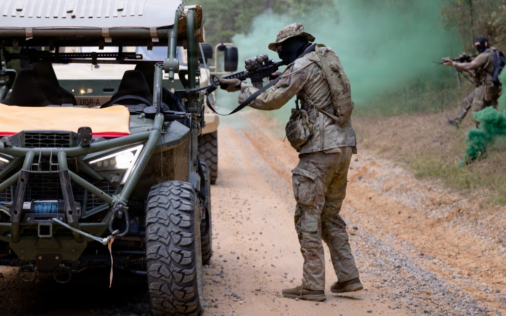 The image depicts what appears to be a military training exercise on a dirt road in a wooded area. Two armed personnel in camouflage uniforms and face coverings are taking tactical positions near military utility vehicles, with green smoke grenades deployed in the background. The scene includes off-road tactical vehicles and suggests a simulated combat or convoy security drill.