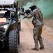 The image depicts what appears to be a military training exercise on a dirt road in a wooded area. Two armed personnel in camouflage uniforms and face coverings are taking tactical positions near military utility vehicles, with green smoke grenades deployed in the background. The scene includes off-road tactical vehicles and suggests a simulated combat or convoy security drill.