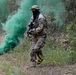 Two armed individuals in full camouflage uniforms and black face coverings are moving through a wooded, overgrown area. A green smoke grenade has been deployed on the ground, producing a dense cloud of green smoke. The scene appears to depict a military training exercise or tactical field operation.