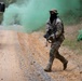 A masked soldier in full camouflage gear walks along a dirt/gravel road during what appears to be a military training exercise. Green smoke grenades are deployed on both sides of the road, creating a tactical smoke screen. A military tactical vehicle (LMTV/FMTV) is visible on the left side of the frame.