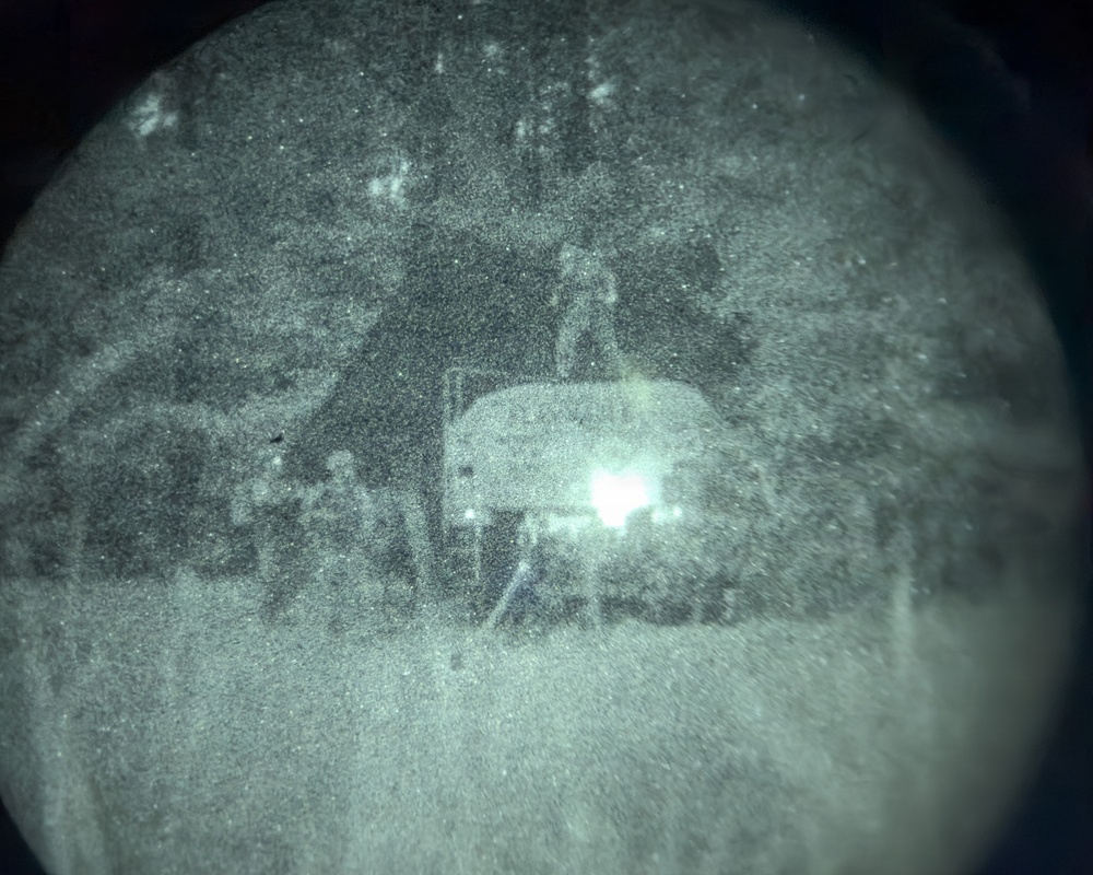 A night vision device capture showing a low-light outdoor scene with what appears to be a vehicle and surrounding vegetation or structures. The circular vignette is characteristic of monocular or binocular night vision optics. Image quality is heavily degraded by grain and low ambient light conditions.