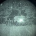 A night vision device capture showing a low-light outdoor scene with what appears to be a vehicle and surrounding vegetation or structures. The circular vignette is characteristic of monocular or binocular night vision optics. Image quality is heavily degraded by grain and low ambient light conditions.