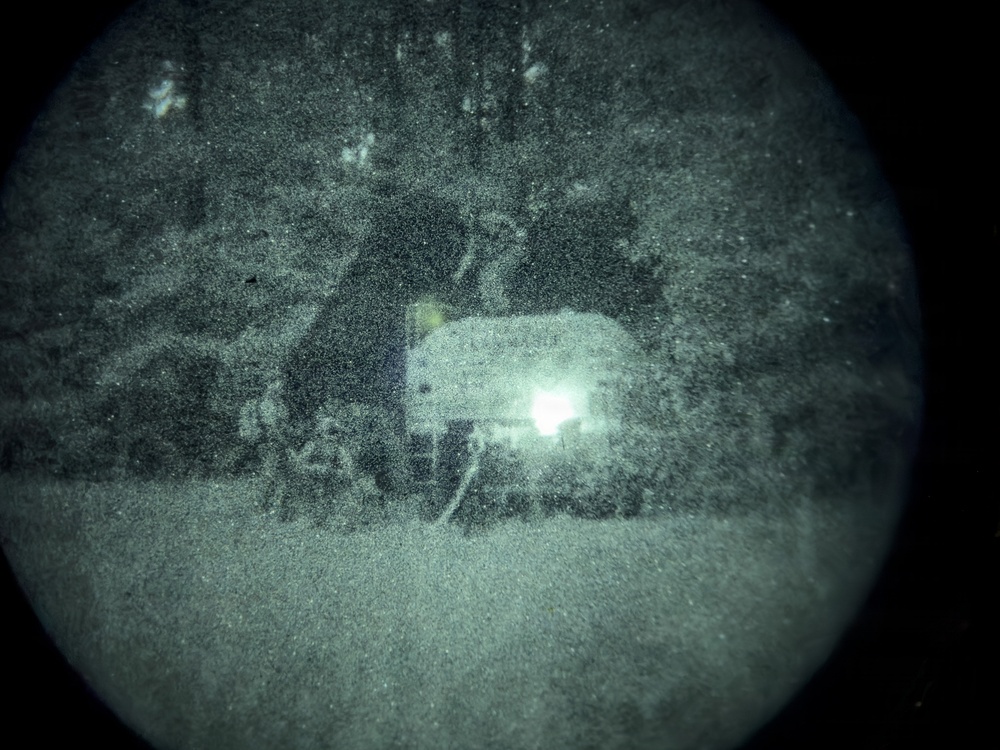 A night vision image captured through a circular optical device, showing what appears to be a vehicle with an active headlight or spotlight in a wooded or outdoor environment. The image is heavily grainy and low-resolution, characteristic of generation-1 or generation-2 night vision optics. A bright light source near the center-right dominates the scene, with dark tree-like shapes visible in the background.