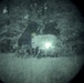 A night vision image captured through a circular optical device, showing what appears to be a vehicle with an active headlight or spotlight in a wooded or outdoor environment. The image is heavily grainy and low-resolution, characteristic of generation-1 or generation-2 night vision optics. A bright light source near the center-right dominates the scene, with dark tree-like shapes visible in the background.