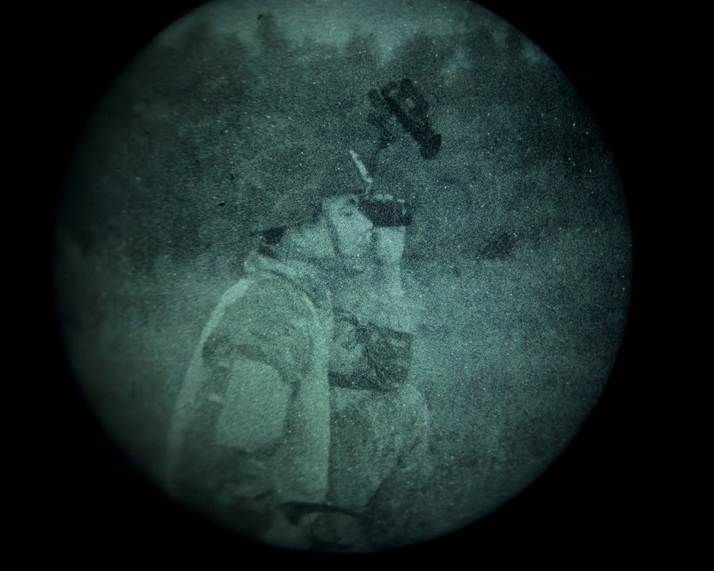 A night vision device capture showing a person wearing military-style tactical gear and a helmet with mounted equipment. The circular field of view and green-tinted grainy appearance are characteristic of night vision optics. The subject appears to be in a profile or three-quarter view orientation outdoors.