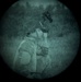 A night vision device capture showing a person wearing military-style tactical gear and a helmet with mounted equipment. The circular field of view and green-tinted grainy appearance are characteristic of night vision optics. The subject appears to be in a profile or three-quarter view orientation outdoors.
