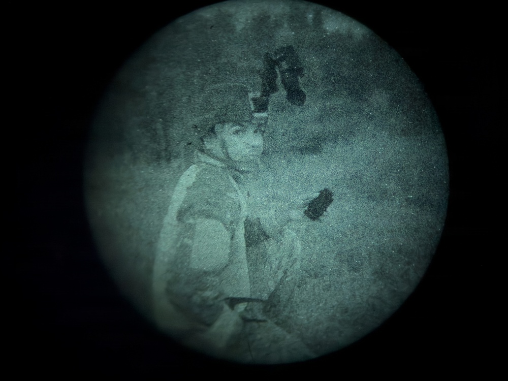 A night vision image captured through a circular lens showing a single individual in what appears to be an outdoor nighttime setting. The green-tinted monochromatic image is characteristic of image intensifier night vision optics. The subject appears to be wearing layered clothing and a helmet or headgear with mounted equipment.