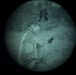 A night vision image captured through a circular lens showing a single individual in what appears to be an outdoor nighttime setting. The green-tinted monochromatic image is characteristic of image intensifier night vision optics. The subject appears to be wearing layered clothing and a helmet or headgear with mounted equipment.