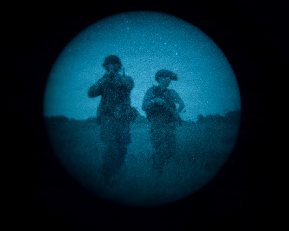 The image is captured through a night-vision or thermal optic device, showing a circular field of view with a blue-tinted monochrome appearance. Two figures in military gear and helmets are visible moving through what appears to be an open field at night. Stars are faintly visible in the sky above the figures.