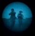 The image is captured through a night-vision or thermal optic device, showing a circular field of view with a blue-tinted monochrome appearance. Two figures in military gear and helmets are visible moving through what appears to be an open field at night. Stars are faintly visible in the sky above the figures.