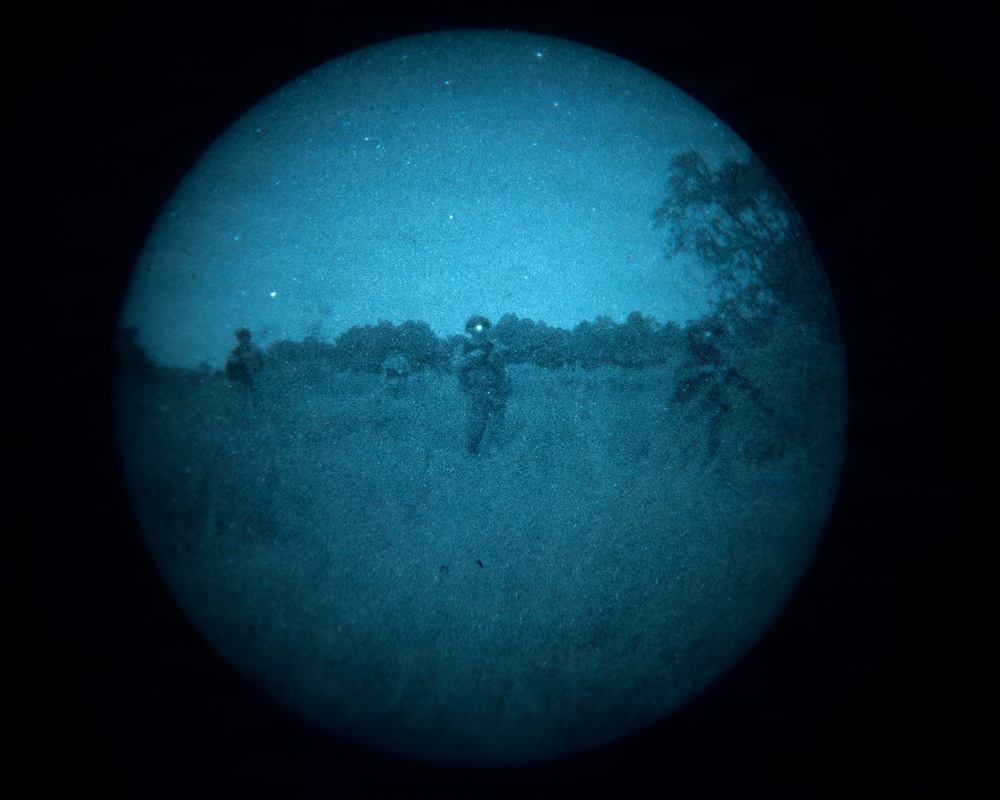 This image appears to be captured through a night vision device, displaying a circular field of view with a characteristic blue-tinted monochrome appearance. Several human figures are visible in an open field or clearing, with tree lines visible in the background. The grainy, low-resolution quality is consistent with image intensifier tube night vision optics.