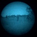 This image appears to be captured through a night vision device, displaying a circular field of view with a characteristic blue-tinted monochrome appearance. Several human figures are visible in an open field or clearing, with tree lines visible in the background. The grainy, low-resolution quality is consistent with image intensifier tube night vision optics.