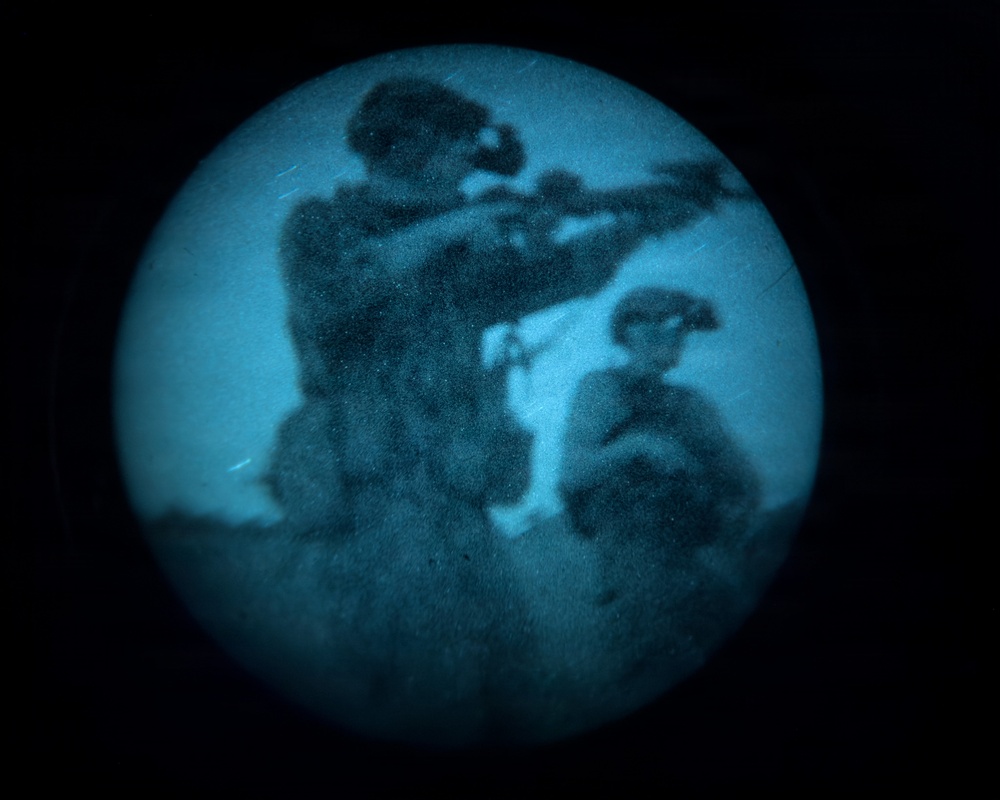 The image appears to be captured through a night vision or thermal imaging device, showing a circular field of view with a blue-green tint characteristic of image intensifier technology. Two figures are visible, with the foreground figure appearing to aim a long weapon such as a rifle. The grainy, low-resolution quality is consistent with night vision optics.
