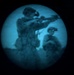 The image appears to be captured through a night vision or thermal imaging device, showing a circular field of view with a blue-green tint characteristic of image intensifier technology. Two figures are visible, with the foreground figure appearing to aim a long weapon such as a rifle. The grainy, low-resolution quality is consistent with night vision optics.