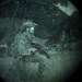 The image is captured through a night vision device, showing a circular green-tinted field of view characteristic of image intensifier optics. A single armed individual in military-style gear and helmet is visible in the center of the frame. The scene appears to be an outdoor nighttime environment with structures or vegetation visible in the background.