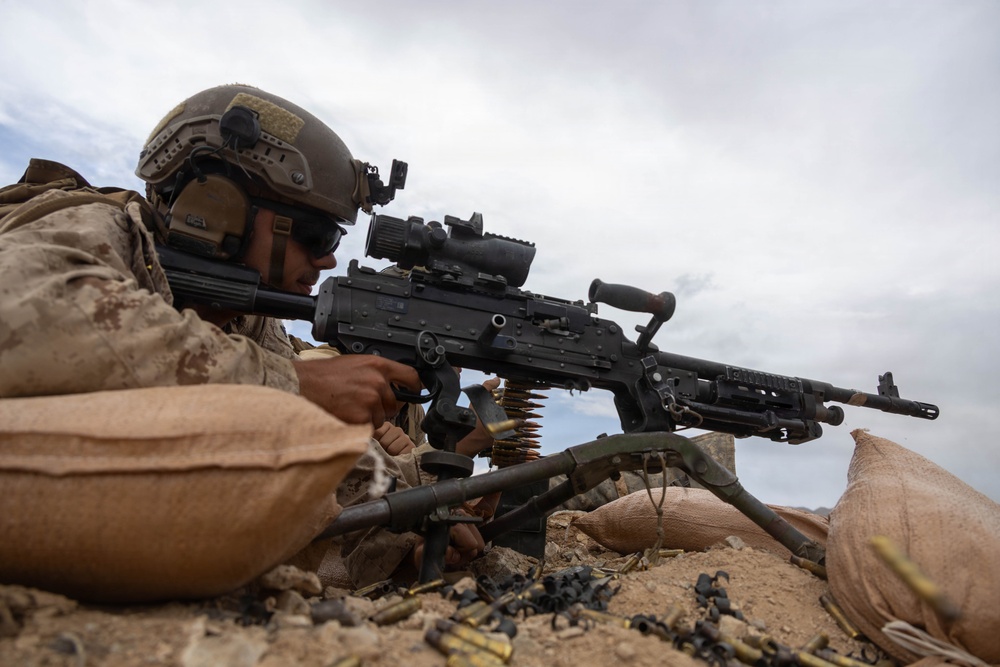 A military service member in desert camouflage uniform is positioned behind a belt-fed machine gun at a fortified fighting position reinforced with sandbags. The soldier is in a prone or low firing position, aiming downrange with spent brass casings visible on the ground. The setting appears to be an arid, desert environment under an overcast sky.