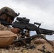 A military service member in desert camouflage uniform is positioned behind a belt-fed machine gun at a fortified fighting position reinforced with sandbags. The soldier is in a prone or low firing position, aiming downrange with spent brass casings visible on the ground. The setting appears to be an arid, desert environment under an overcast sky.