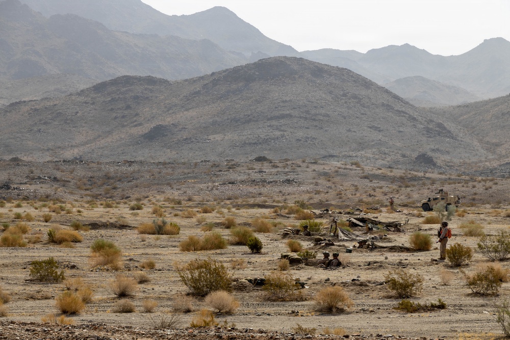 A desert landscape showing what appears to be a military field exercise or training operation, with personnel and equipment positioned in an arid, scrub-covered terrain. Rocky mountains rise in the background under a hazy sky. A small forward position or temporary camp is visible in the middle distance, with at least one armored vehicle and several personnel present.