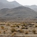 A desert landscape showing what appears to be a military field exercise or training operation, with personnel and equipment positioned in an arid, scrub-covered terrain. Rocky mountains rise in the background under a hazy sky. A small forward position or temporary camp is visible in the middle distance, with at least one armored vehicle and several personnel present.