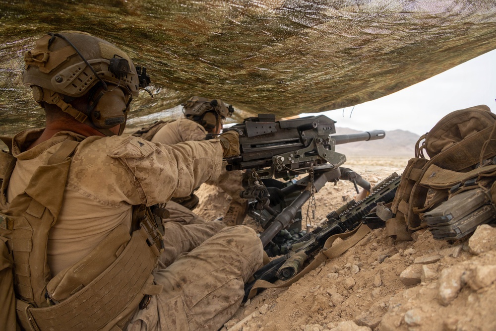 Two military personnel in desert camouflage uniforms are positioned under a camouflage netting in an arid, rocky terrain. They are operating what appears to be an Mk 19 grenade launcher mounted on a tripod, with an M4-style rifle also visible in the foreground. The setting suggests a training exercise or combat operation in a desert environment.