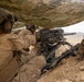 Two military personnel in desert camouflage uniforms are positioned under a camouflage netting in an arid, rocky terrain. They are operating what appears to be an Mk 19 grenade launcher mounted on a tripod, with an M4-style rifle also visible in the foreground. The setting suggests a training exercise or combat operation in a desert environment.