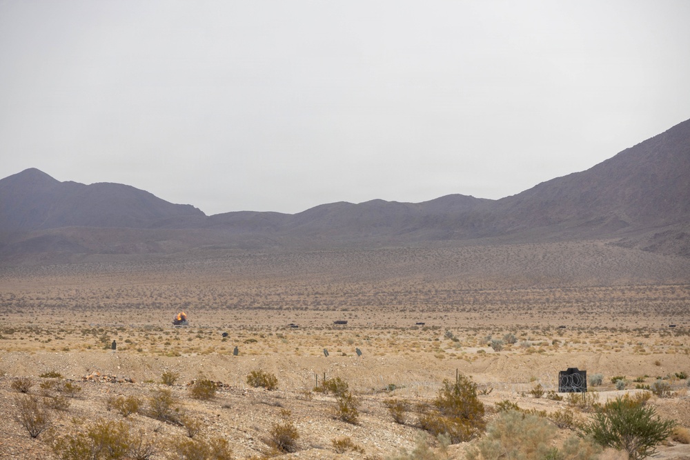 A wide desert landscape with arid terrain, sparse scrub vegetation, and mountain ranges in the background under an overcast sky. In the mid-ground, a small orange fireball or explosion is visible, suggesting a detonation or controlled blast event. Several structures or objects are scattered across the desert floor, consistent with a military training range or test site.