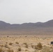 A wide desert landscape with arid terrain, sparse scrub vegetation, and mountain ranges in the background under an overcast sky. In the mid-ground, a small orange fireball or explosion is visible, suggesting a detonation or controlled blast event. Several structures or objects are scattered across the desert floor, consistent with a military training range or test site.