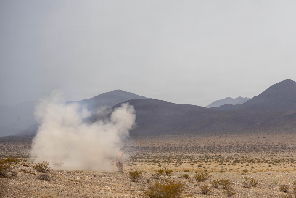 A desert landscape with a large plume of white smoke rising from the ground, suggesting a detonation, flare, or smoke grenade deployment. The terrain is arid and flat with sparse desert scrub vegetation, backed by dark mountain ridges under a hazy, overcast sky. A small human figure is partially visible at the base of the smoke cloud.
