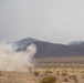 A desert landscape with a large plume of white smoke rising from the ground, suggesting a detonation, flare, or smoke grenade deployment. The terrain is arid and flat with sparse desert scrub vegetation, backed by dark mountain ridges under a hazy, overcast sky. A small human figure is partially visible at the base of the smoke cloud.