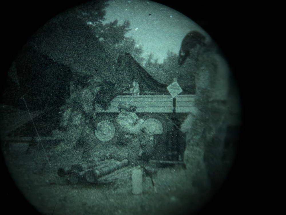 A heavily degraded, circular-vignette image that appears to be viewed through a lens or scope, rendered in a teal/green monochromatic tone. The scene depicts what appears to be an outdoor setting with a tent, trees, a motorcycle or small vehicle, and at least one human figure. The extreme grain, distortion, and circular framing significantly reduce image clarity.