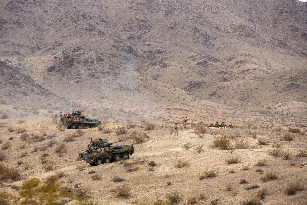 The image shows a military operation in an arid, mountainous desert terrain with two wheeled armored vehicles and several dismounted personnel. The vehicles appear to be Light Armored Vehicles (LAVs) or similar 8-wheeled armored personnel carriers in desert camouflage. Personnel are dispersed across the hillside in what appears to be a training exercise or tactical movement.