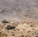 The image shows a military operation in an arid, mountainous desert terrain with two wheeled armored vehicles and several dismounted personnel. The vehicles appear to be Light Armored Vehicles (LAVs) or similar 8-wheeled armored personnel carriers in desert camouflage. Personnel are dispersed across the hillside in what appears to be a training exercise or tactical movement.