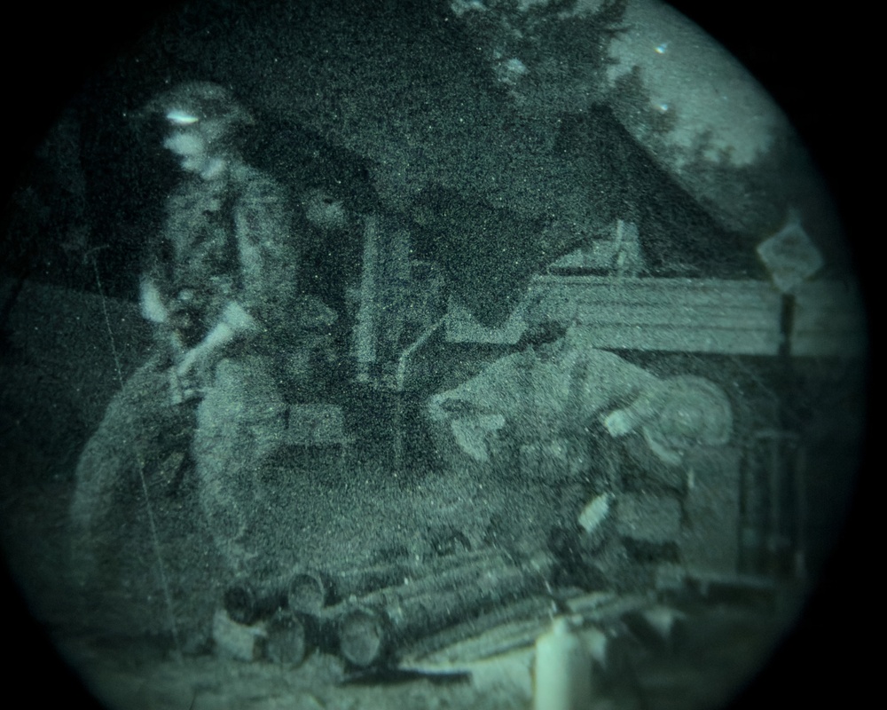 A night vision image showing what appears to be one or more individuals in an outdoor setting near a structure. The circular vignette and green-tinted grainy appearance are characteristic of night vision optics. The scene includes what appears to be equipment or machinery in the foreground.