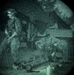 A night vision image showing what appears to be one or more individuals in an outdoor setting near a structure. The circular vignette and green-tinted grainy appearance are characteristic of night vision optics. The scene includes what appears to be equipment or machinery in the foreground.