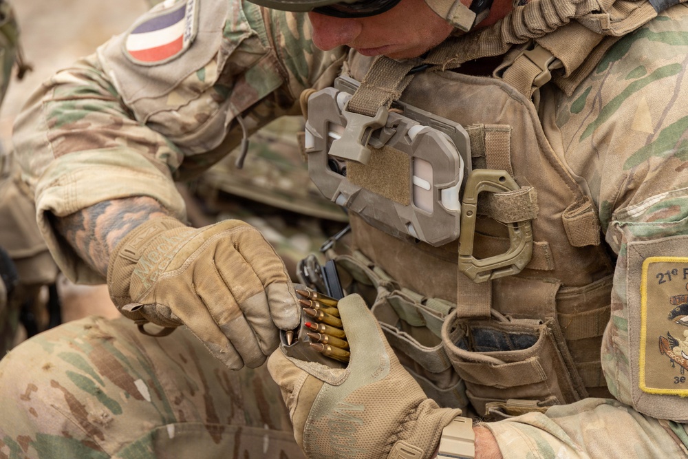 A soldier in multicam camouflage uniform is loading ammunition into a magazine during what appears to be a training or operational exercise. The soldier wears tactical gear including a plate carrier, gloves, and helmet, with a flag patch and unit patch visible on the uniform. The close-up shot focuses on the hands manipulating cartridges and a magazine.