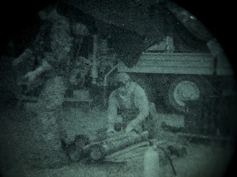 A night-vision or thermal imaging capture showing a person crouched or seated near what appears to be a vehicle or heavy machinery in an outdoor or industrial setting. The circular vignette is characteristic of optics such as binoculars, night-vision goggles, or a scope. Image quality is heavily degraded by grain and low-light conditions.