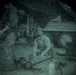 A night-vision or thermal imaging capture showing a person crouched or seated near what appears to be a vehicle or heavy machinery in an outdoor or industrial setting. The circular vignette is characteristic of optics such as binoculars, night-vision goggles, or a scope. Image quality is heavily degraded by grain and low-light conditions.