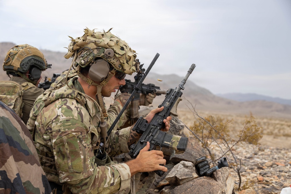 Two armed soldiers in multicam camouflage uniforms are positioned behind rocky cover in an arid desert environment. The foreground soldier is handling/inspecting a rifle while the background soldier appears to be actively firing their weapon, with a visible brass casing being ejected. The setting and equipment suggest a military training exercise or combat operation in a desert region.