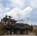 A wheeled armored fighting vehicle, consistent with an LAV-25 or similar 8-wheeled light armored vehicle, is firing its main cannon in a desert training environment. The vehicle is draped in camouflage netting and equipped with radio antennas, with a crew member visible in the commander's hatch. Smoke from the cannon discharge is visible behind the vehicle against a partly cloudy sky.