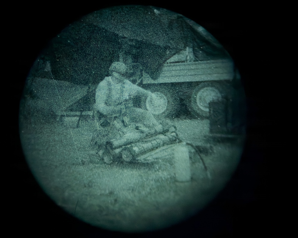 The image appears to be captured through a night vision or thermal optic device, indicated by the circular vignette and green-tinted monochromatic appearance. A figure in military or tactical gear is visible in a crouched or kneeling position near what appears to be a vehicle and tent or tarp structure. The image is grainy and low resolution, consistent with night vision optics.
