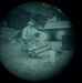 The image appears to be captured through a night vision or thermal optic device, indicated by the circular vignette and green-tinted monochromatic appearance. A figure in military or tactical gear is visible in a crouched or kneeling position near what appears to be a vehicle and tent or tarp structure. The image is grainy and low resolution, consistent with night vision optics.