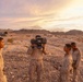 Four military personnel in desert camouflage uniforms are gathered in an arid, rocky desert environment during golden hour. One soldier is shouldering and aiming what appears to be a man-portable missile launcher or similar crew-served weapon system. Military trucks are visible in the background against a mountain range.