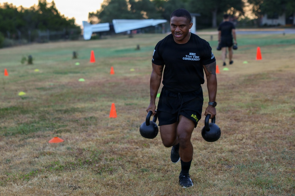 A soldier in U.S. Army physical training gear carries two kettlebells across a grass field marked with orange and yellow cones, performing what appears to be a farmer's carry exercise. The text on his shirt reads 'HHC 71st T100 COMMANDER.' A second individual in black clothing is visible in the background performing a similar activity.