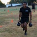 A soldier in U.S. Army physical training gear carries two kettlebells across a grass field marked with orange and yellow cones, performing what appears to be a farmer's carry exercise. The text on his shirt reads 'HHC 71st T100 COMMANDER.' A second individual in black clothing is visible in the background performing a similar activity.