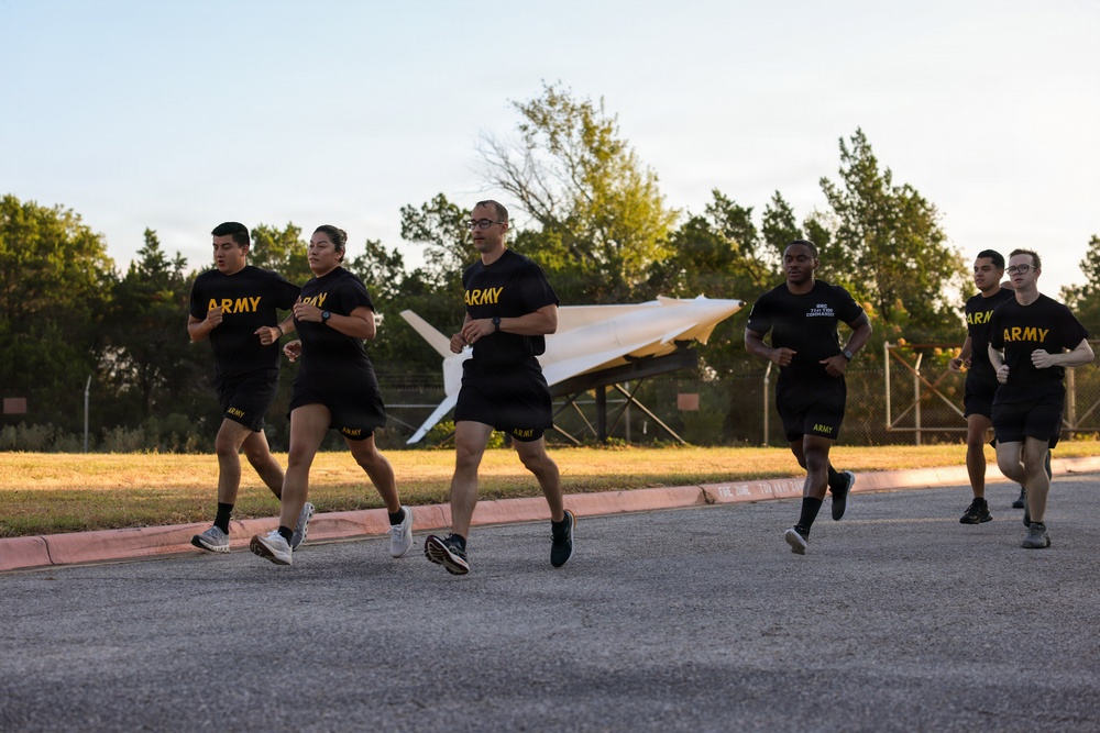 A group of U.S. Army soldiers in physical training (PT) uniforms are running together on a paved road at what appears to be a military installation. The soldiers are wearing matching black Army PT shirts and shorts with gold lettering. In the background, a static display of what appears to be a missile or rocket is visible behind a fence.