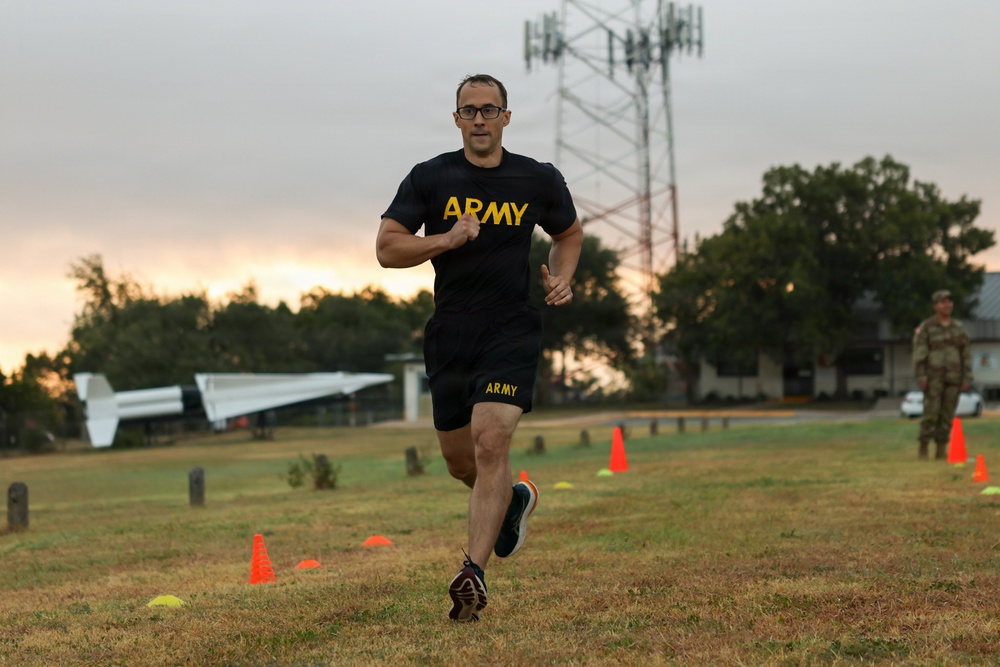 A male soldier wearing U.S. Army physical training (PT) uniform — black shirt and shorts with gold 'ARMY' lettering — is running across a grass field during what appears to be an early morning fitness test or training event. Orange and yellow cones mark a course on the field, and a second soldier in camouflage uniform observes in the background. A static aircraft display and a communications tower are visible in the background, suggesting a military installation.