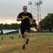 A male soldier wearing U.S. Army physical training (PT) uniform — black shirt and shorts with gold 'ARMY' lettering — is running across a grass field during what appears to be an early morning fitness test or training event. Orange and yellow cones mark a course on the field, and a second soldier in camouflage uniform observes in the background. A static aircraft display and a communications tower are visible in the background, suggesting a military installation.