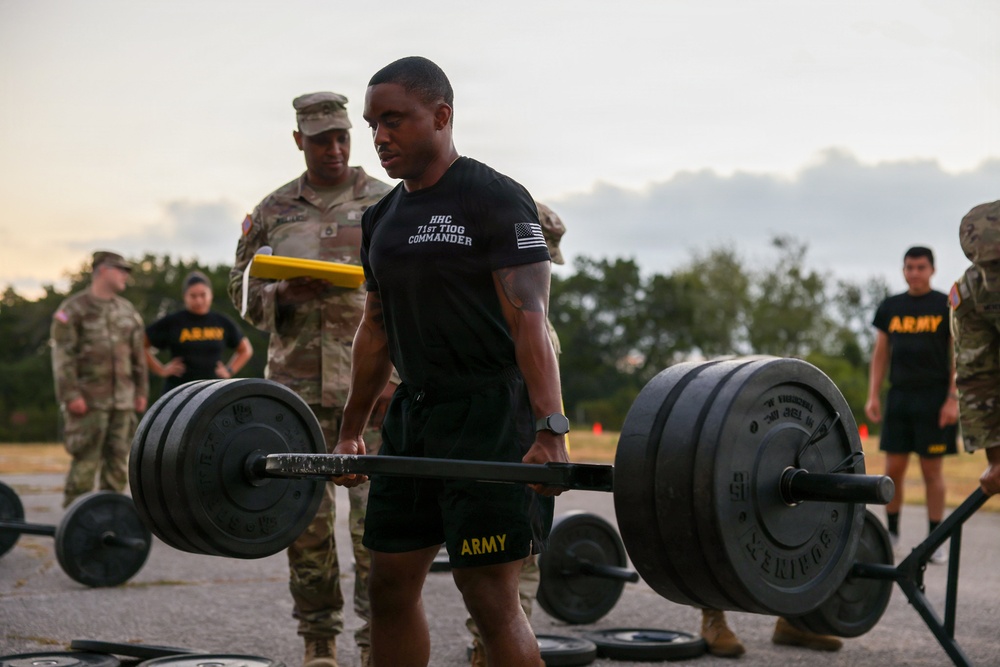 A U.S. Army soldier wearing a black 'HHC 71st TIOG Commander' shirt performs a deadlift with a heavily loaded barbell during what appears to be an Army Combat Fitness Test (ACFT) or physical fitness evaluation. Several other soldiers in Army PT uniforms and one in Army Combat Uniform (ACU) with a clipboard observe and score the event. The setting is an outdoor paved area during early morning or evening light.