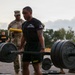 A U.S. Army soldier wearing a black 'HHC 71st TIOG Commander' shirt performs a deadlift with a heavily loaded barbell during what appears to be an Army Combat Fitness Test (ACFT) or physical fitness evaluation. Several other soldiers in Army PT uniforms and one in Army Combat Uniform (ACU) with a clipboard observe and score the event. The setting is an outdoor paved area during early morning or evening light.