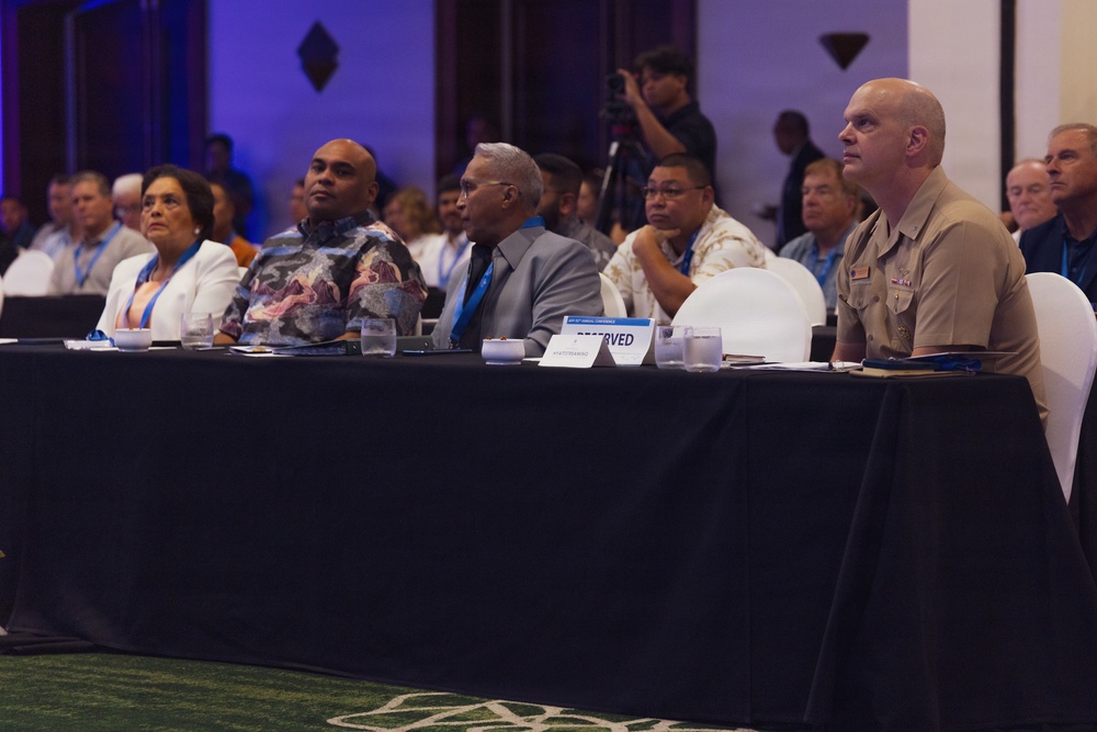 A formal conference or summit is taking place in a large ballroom or event hall. Several attendees are seated at a front-row table with a black tablecloth, including a military officer in tan uniform and civilians wearing blue lanyards. A photographer is visible in the background capturing the event.